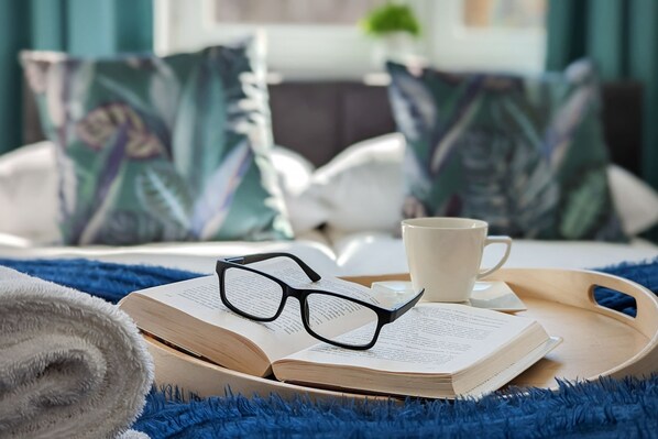 A cozy reading setup with an open book, glasses, and a cup on a tray, surrounded by a blue blanket and pillows.