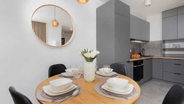 Un comedor moderno con una mesa redonda de madera dispuesta para cuatro comensales, un jarrón blanco con flores y un espejo grande en la pared.