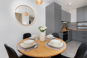 A modern dining area with a round wooden table set for four, a white vase with flowers, and a large mirror on the wall.