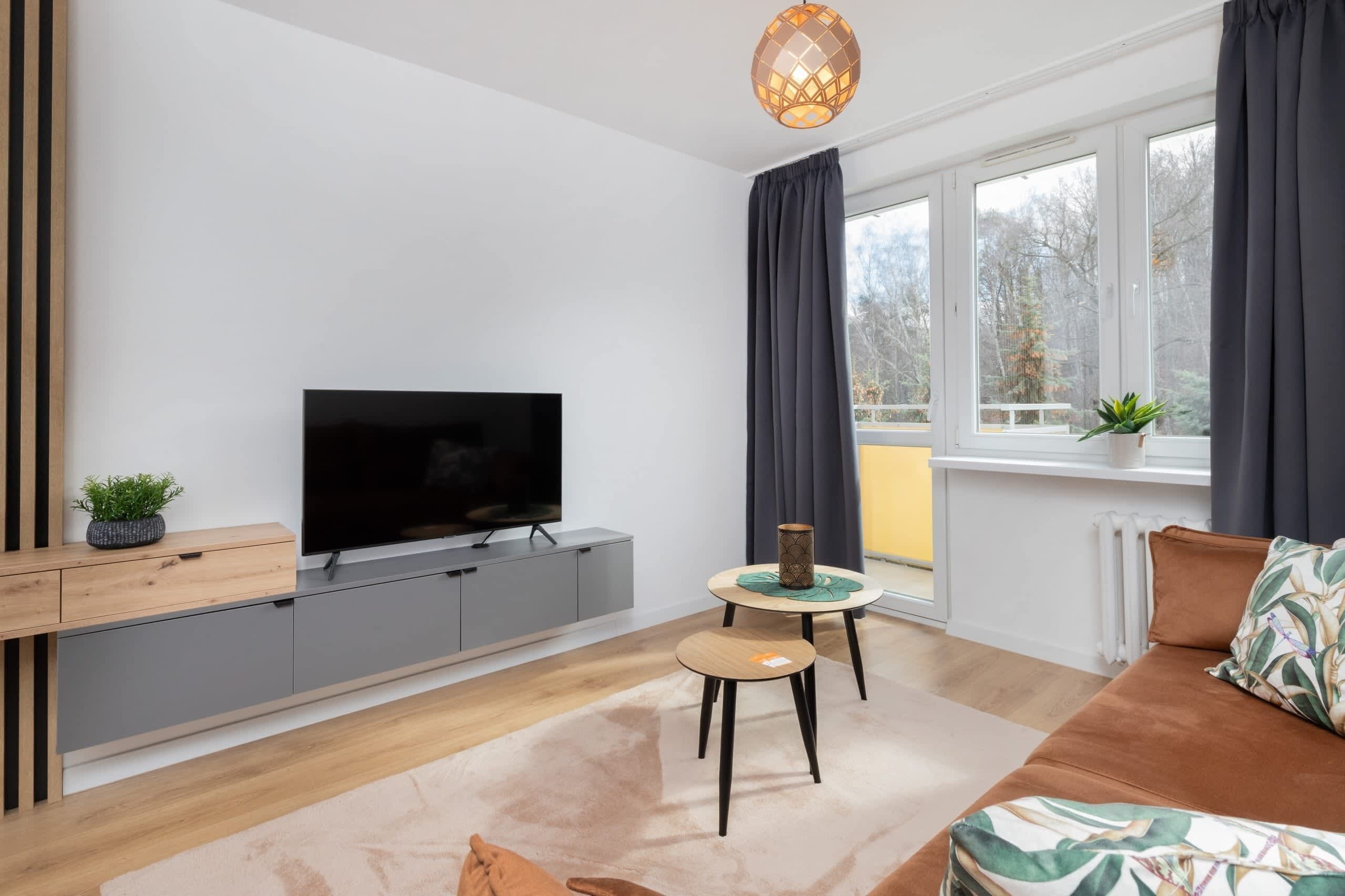 A modern living room with a flat-screen TV, grey cabinets, a round wooden table, a brown sofa, and a large window with curtains.