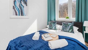 Un dormitorio moderno con una cama grande, cortinas color turquesa, una ventana con vistas, un cuadro abstracto enmarcado y una manta azul con textura.