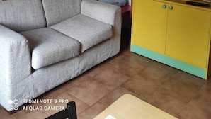 Una sala de estar con un sofá gris, una estantería amarilla, un cartel en la pared y un libro rojo sobre el sofá.