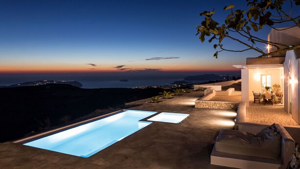 A modern outdoor pool area with a view of the sea at dusk, a patio with a table and chairs, and a cozy seating area.