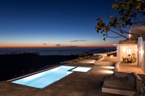 Un área moderna de piscina al aire libre con vista al mar al anochecer, un patio con mesa y sillas, y un rincón de descanso acogedor.