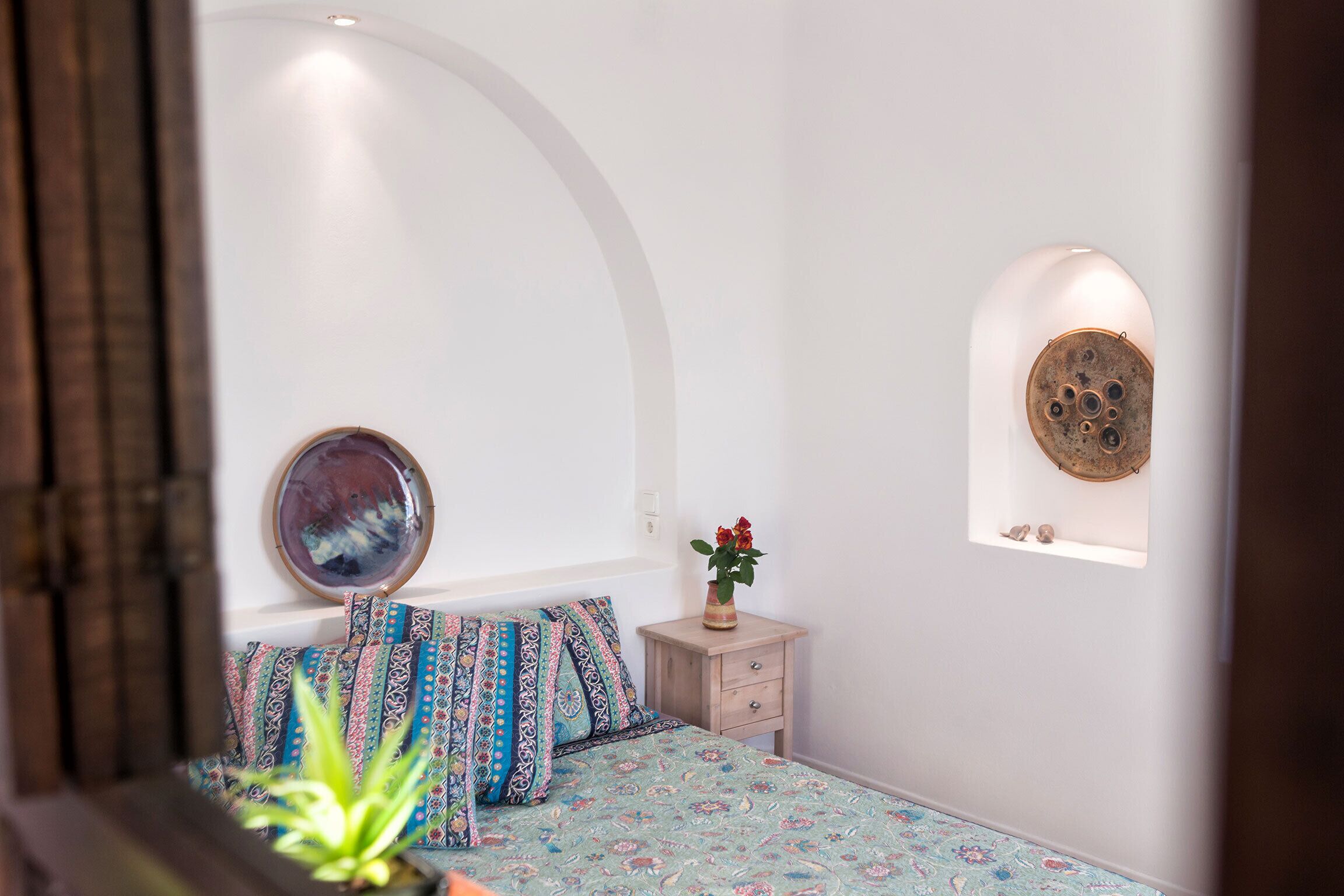 Un dormitorio con cama, almohadas estampadas, una mesita de noche con un florero, un espejo en la pared y un estante con adornos.