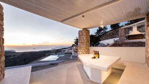 A modern outdoor seating area with a stone pillar, a white table, and a view of the sea at sunset.
