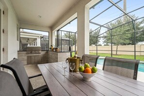 A covered patio with a dining table, chairs, a grill, and a view of a swimming pool.