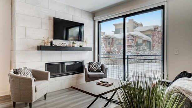 A modern living room with a fireplace, a flat-screen TV mounted on the wall, and a large window offering a view of snow-covered buildings.