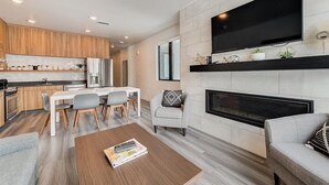 A modern living room with a fireplace, a flat-screen TV mounted above it, and a dining area with a white table and chairs.