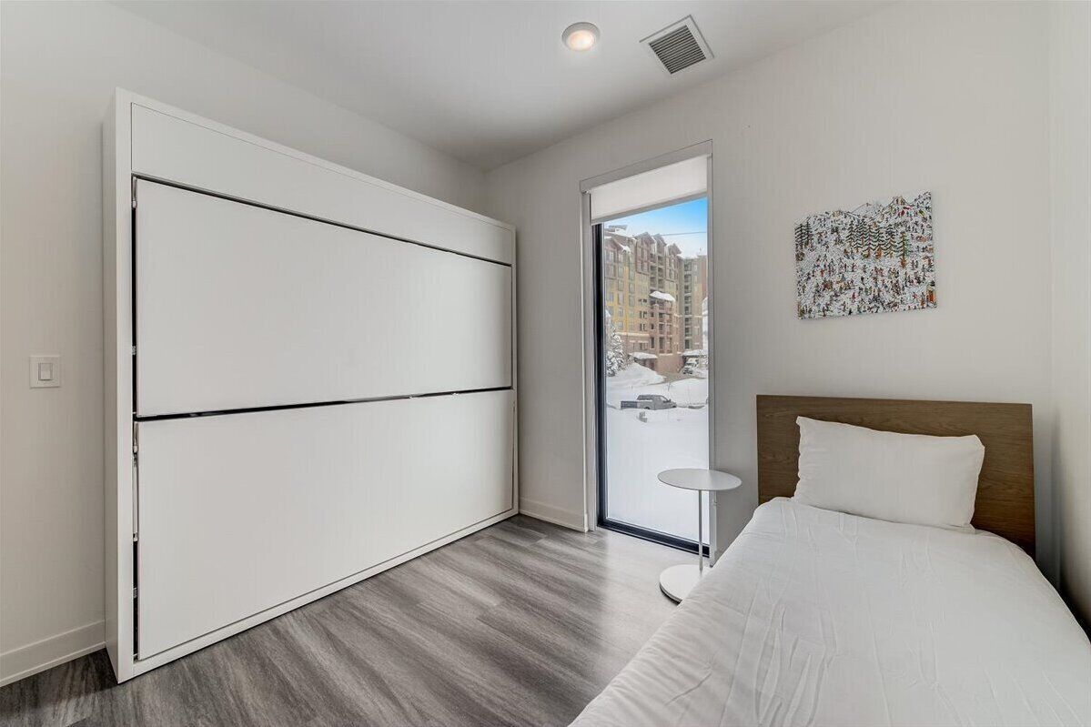 A modern hotel room with a large sliding door, a bed with white bedding, a small round table, and a cityscape view through the window.