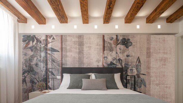 A bedroom with a bed, wooden beams, a headboard, and a floral wallpaper.