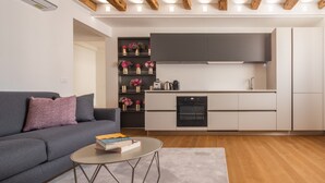 A modern living room with a grey sofa, a coffee table with books, a kitchen area with grey cabinets, and wooden beams on the ceiling. - Ruga Bella Gold Apartment R&R (Venecia)