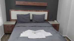 Ein ordentlich bezogenes Bett mit einer gestreiften Tagesdecke, zwei Kissen und einem weißen Handtuch. Auf jeis Nachttisch stehen ein Telefon und eine Lampe. Der Raum hat eine dunkle Wand und ein hölzernes Kopfteil.