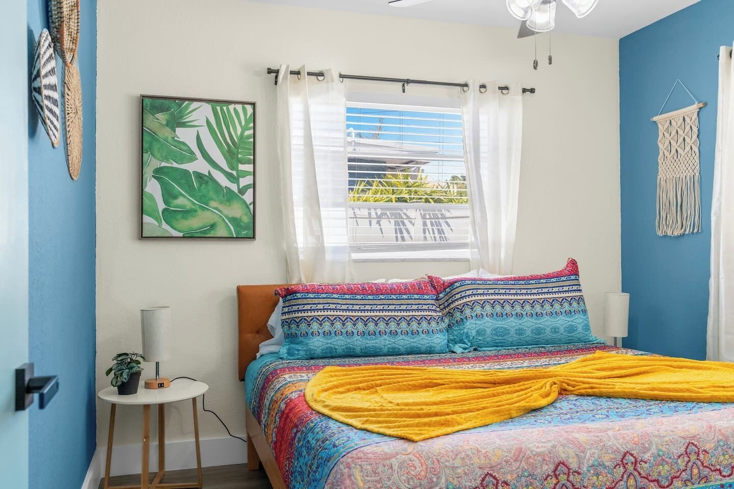 Quarto com cama, janela com cortinas, uma planta e um detalhe em macramê na parede.