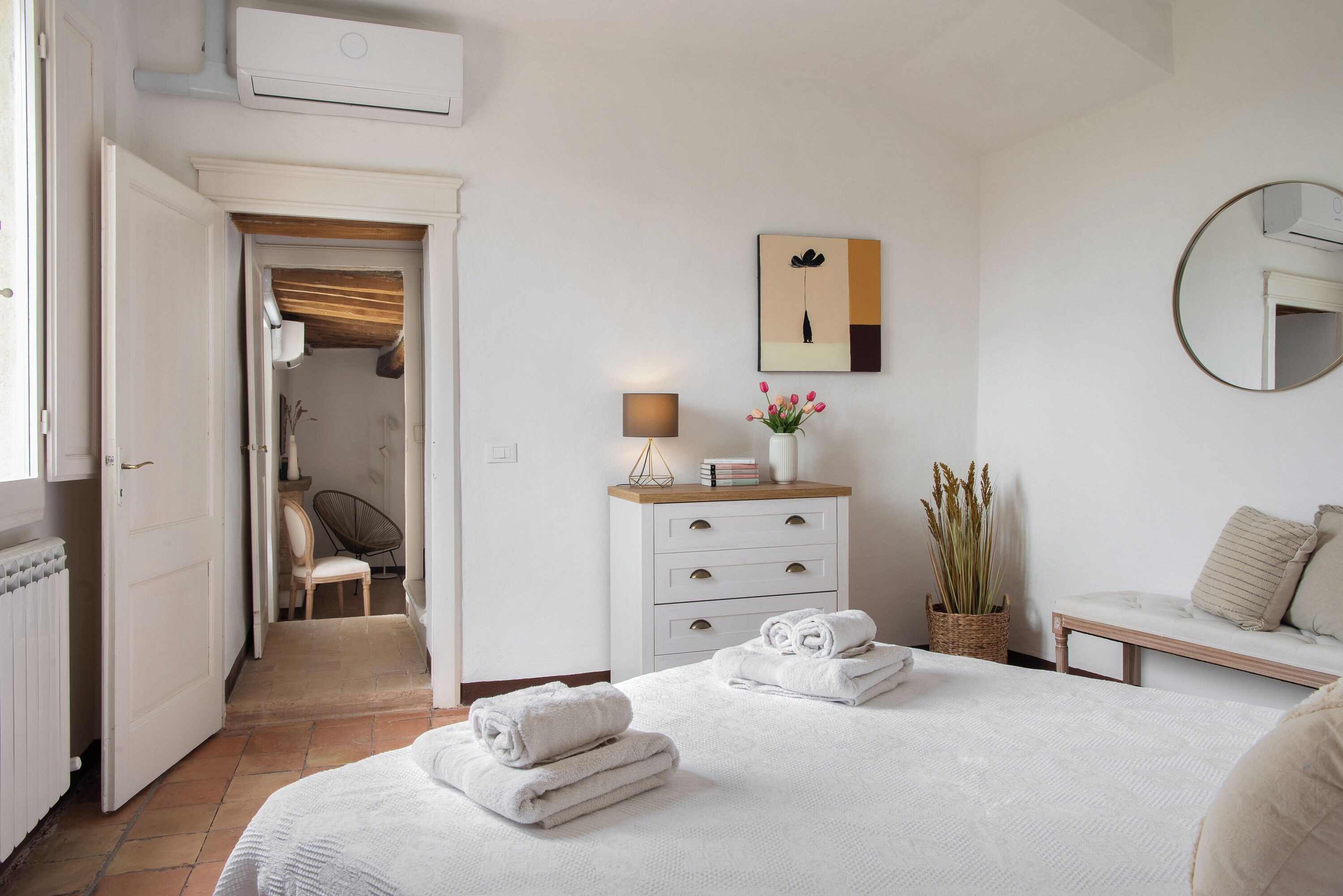 A bedroom with a bed, a bedside table, a lamp, a mirror, a chair, a dresser, and a door leading to another room.
