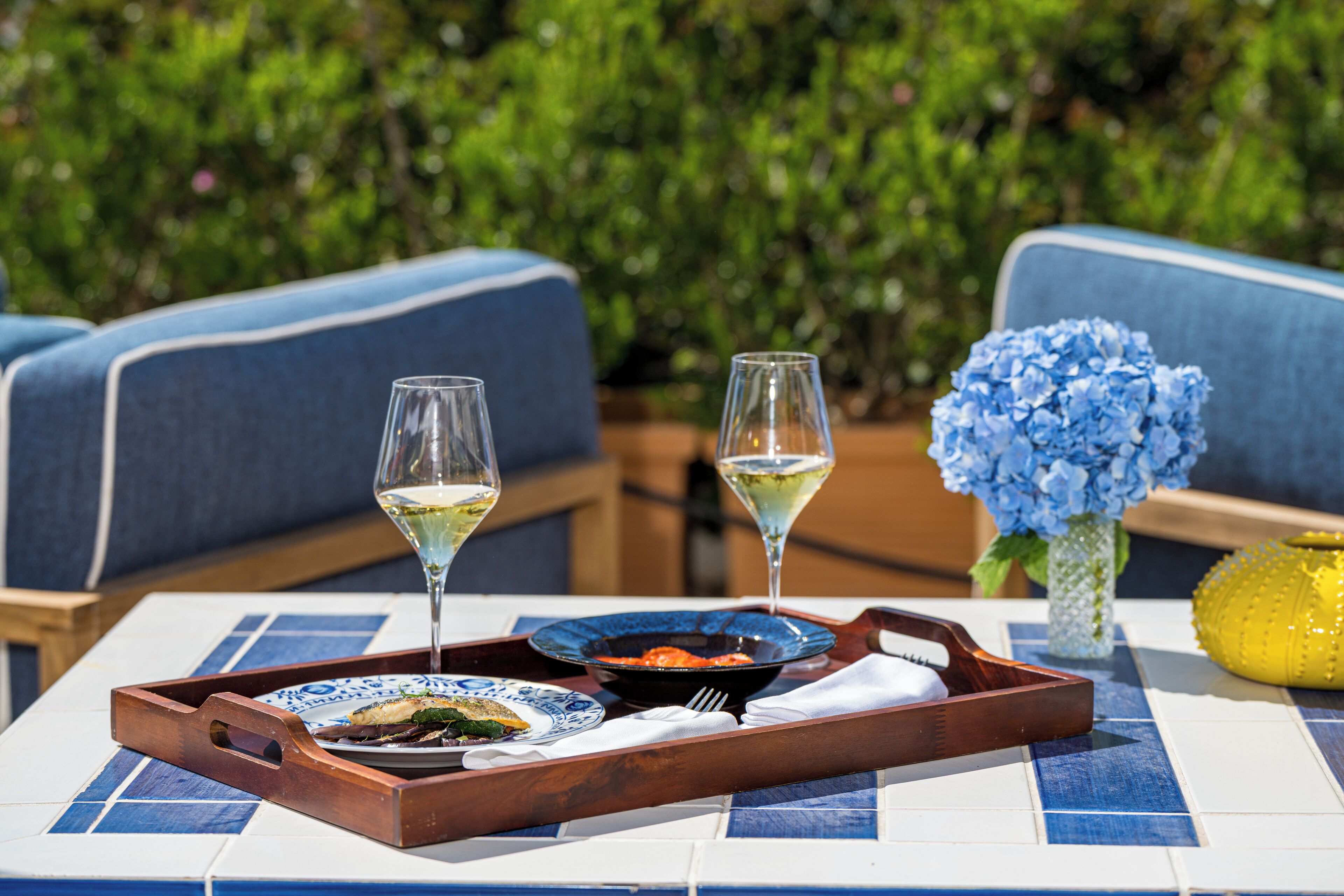 Une table à manger avec un plateau, deux verres de vin blanc, un bol bleu contenant de la nourriture et un vase rempli de fleurs bleues.