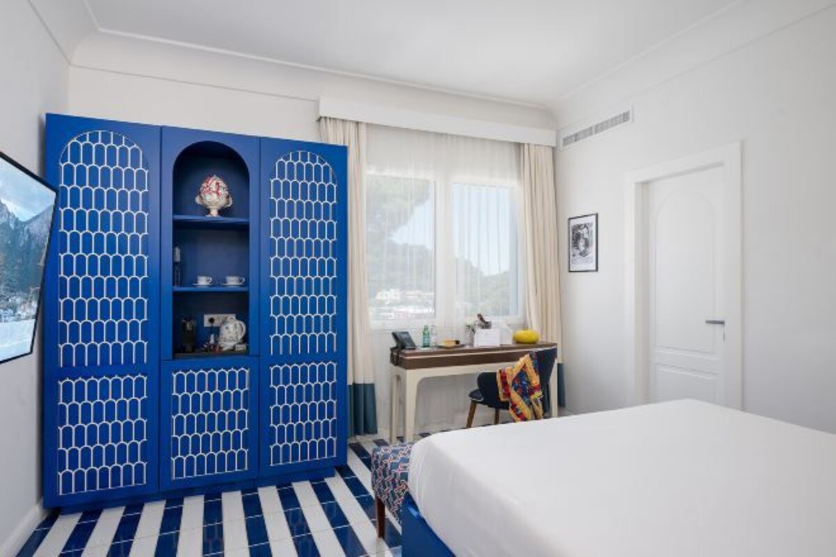 A hotel room with a bed, a blue wardrobe, a desk with a chair, and a window with curtains.