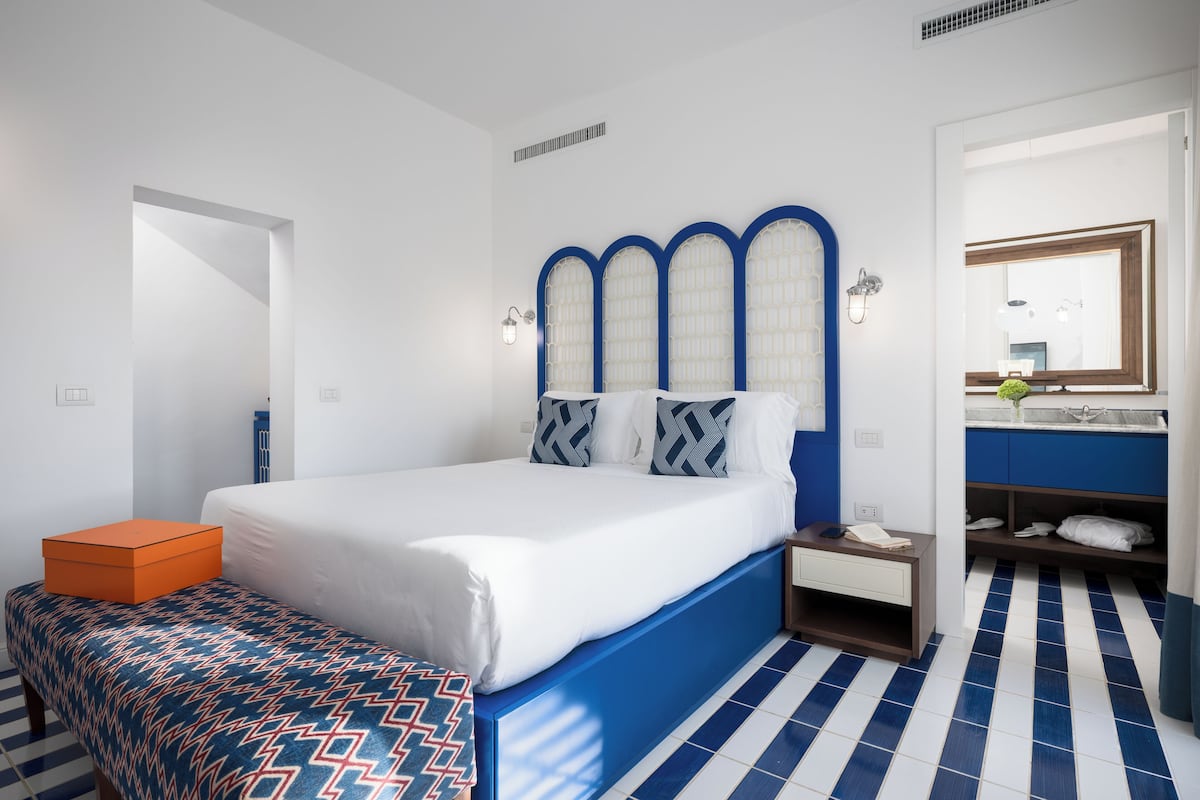Une chambre à coucher comprenant un lit, une table de chevet, un banc, une salle de bain avec un lavabo et un miroir, et un sol carrelé.