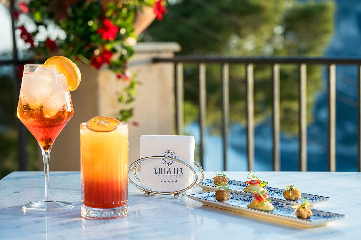 Une table avec un verre à martini, un grand verre de jus d’orange, une pancarte indiquant « Villa Lià Hotel Capri », et une assiette de hors-d’œuvre.