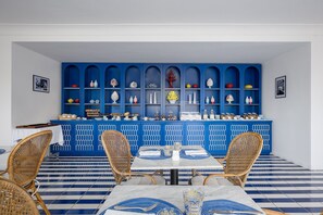 A dining area with a blue and white tiled floor, wicker chairs, a white table with blue placemats, and a blue cabinet with shelves of decorative items. - Villa Lia Hotel Capri (Capri)