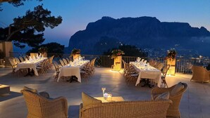 Un área de comedor al aire libre con mesas y sillas de mimbre, con montañas al fondo.