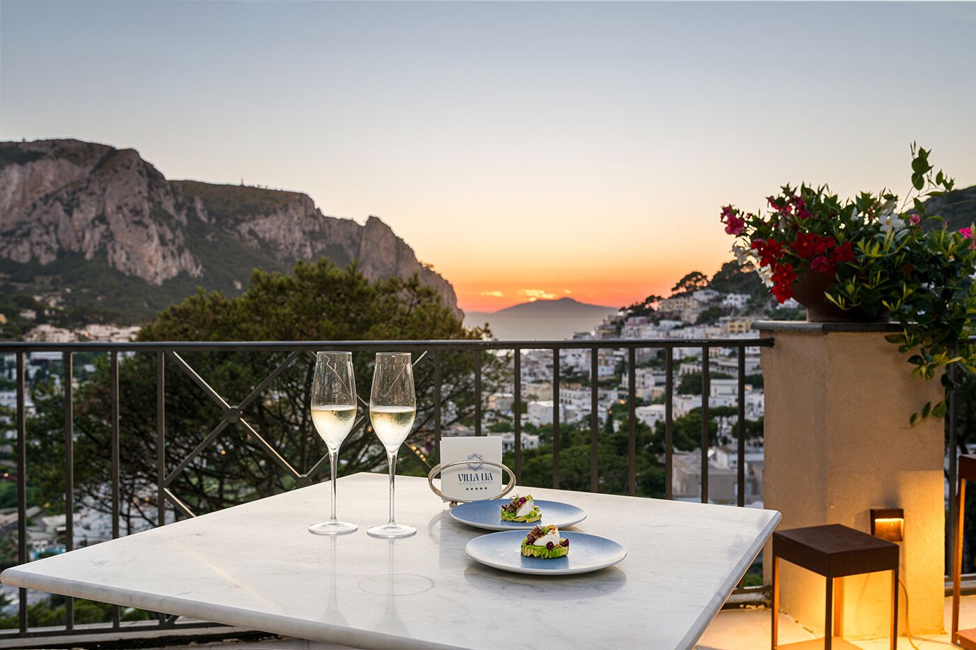 A table with two wine glasses and a menu, overlooking a coastal cityscape at sunset.