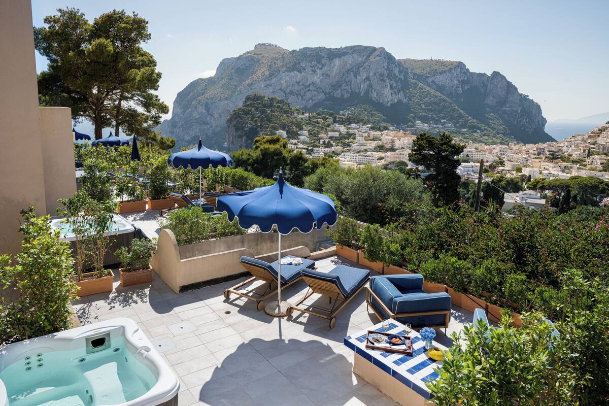 Une terrasse sur le toit, dotée de chaises longues, d’un jacuzzi et de parasols, offrant une vue sur une ville côtière et une montagne.