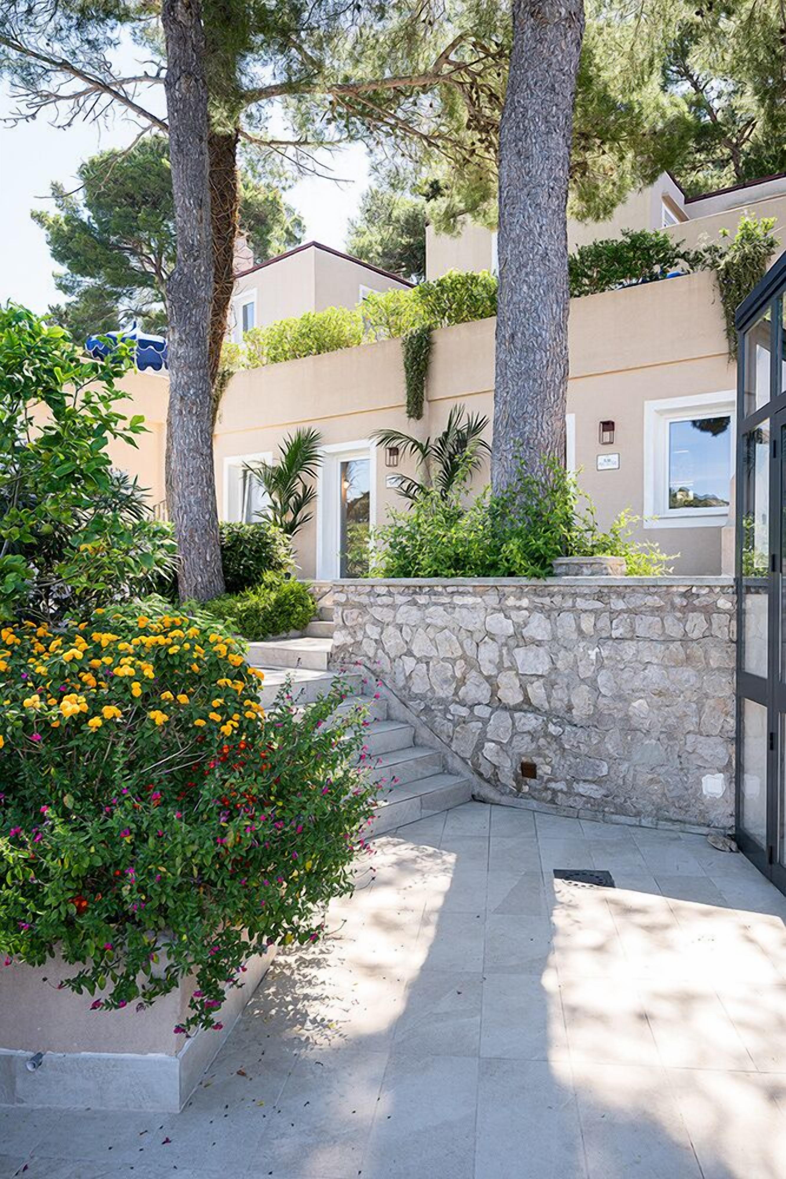 A courtyard with stone walls, a variety of plants, and a building with a beige facade and large windows.