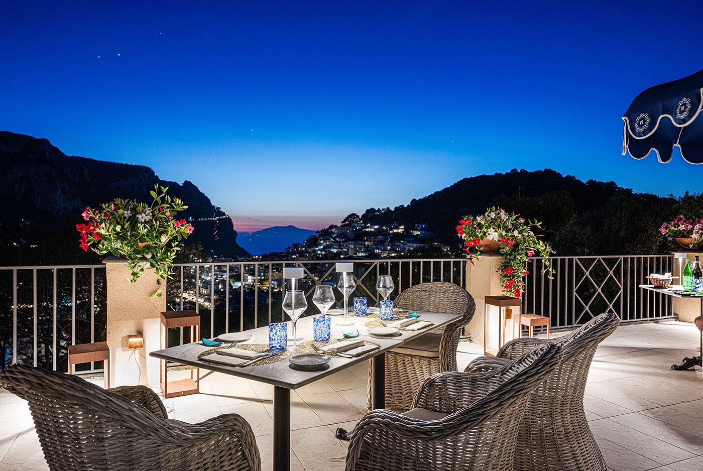 An outdoor dining area with wicker furniture, a table set for two, and flower arrangements.