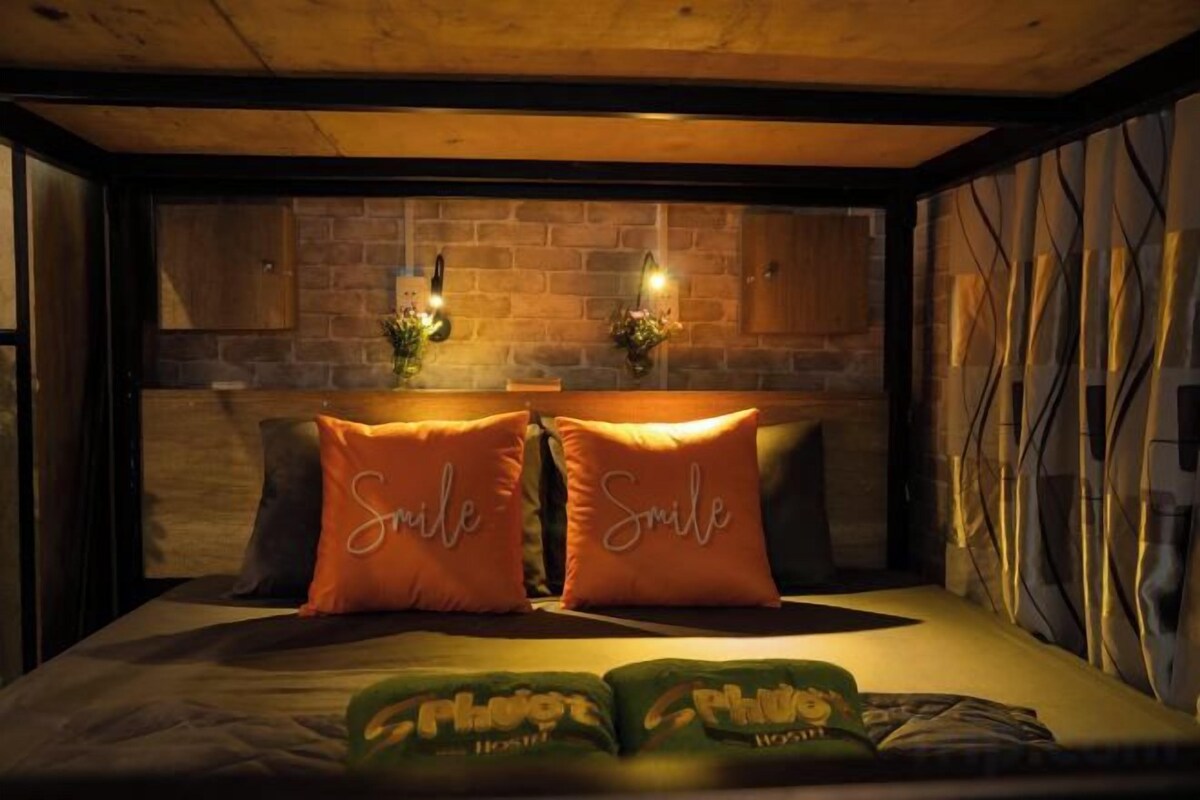 Una cama con dos almohadas anaranjadas, un plaid verde y una pared de ladrillo con dos luces fijadas a la pared.