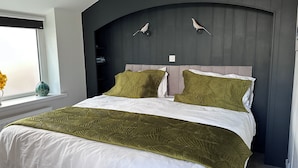 A bedroom with a bed, two wall-mounted bird sculptures, a window with a view, and a shelf with books. - The Nook (Criccieth)