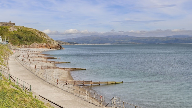 A coastal walkway with a series of wooden piers extending into the sea, a grassy hill with a structure on top, and a clear sky. - The Nook (Criccieth)