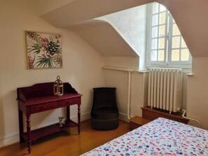 Une chambre avec un plafond en pente, un lit recouvert d’une couvre-lit à motifs floraux, une commode rouge, une chaise noire et un radiateur.