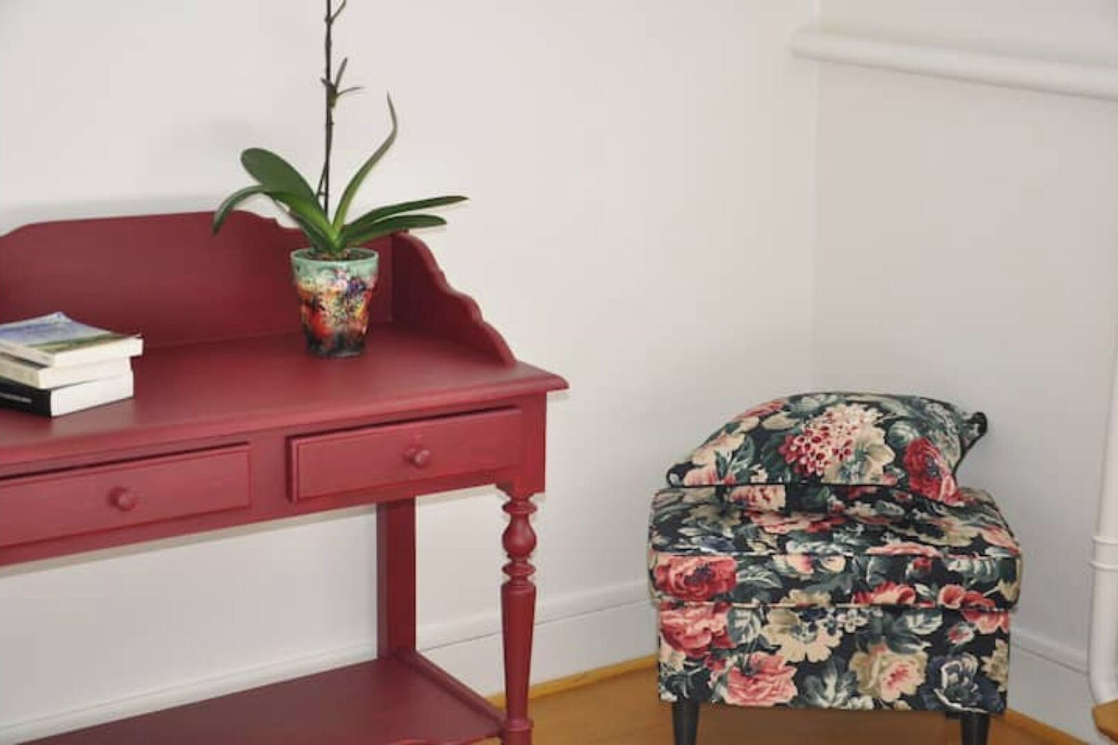 Un escritorio rojo con una planta y libros, un sillón estampado y una pared blanca.