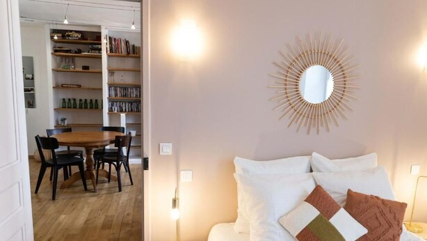 A bedroom with a bed, pillows, a bedside table, a wall-mounted mirror, and a bookshelf in the background. - Peace and comfort in the heart of the city (Draguignan)