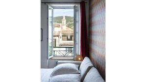 A bedroom with a bed, a window with a view of buildings, and a patterned wall. - Peace and comfort in the heart of the city (Draguignan)