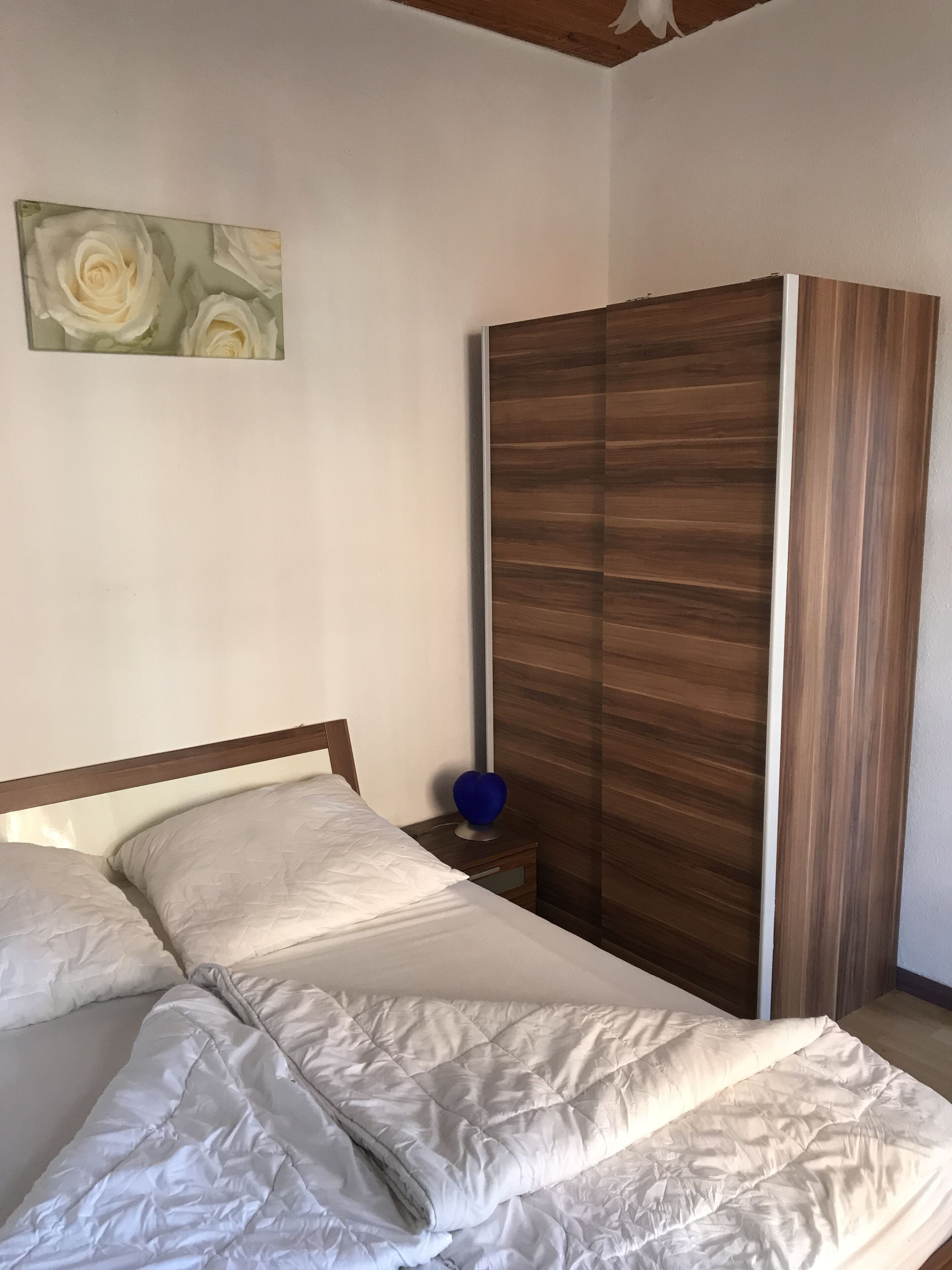 A bedroom with a bed, a nightstand, a blue vase, and a wooden wardrobe.