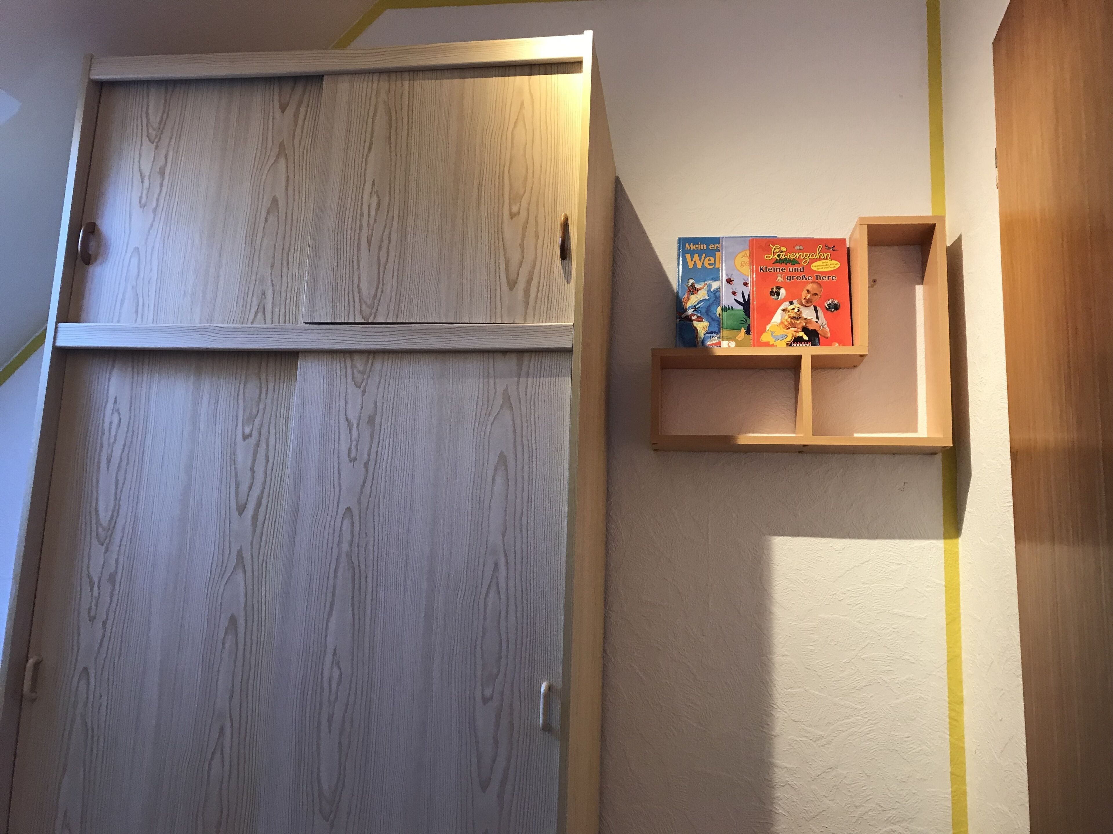 A wooden wardrobe with a sliding door, a small shelf with books, and a yellow line on the wall.