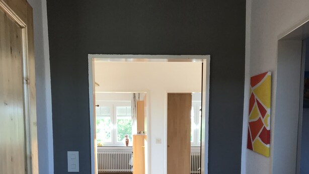 Ein Korridor mit Holzboden, einem grünen Besen und einer Tür zu einem Raum mit Heizkörper und Fenster.