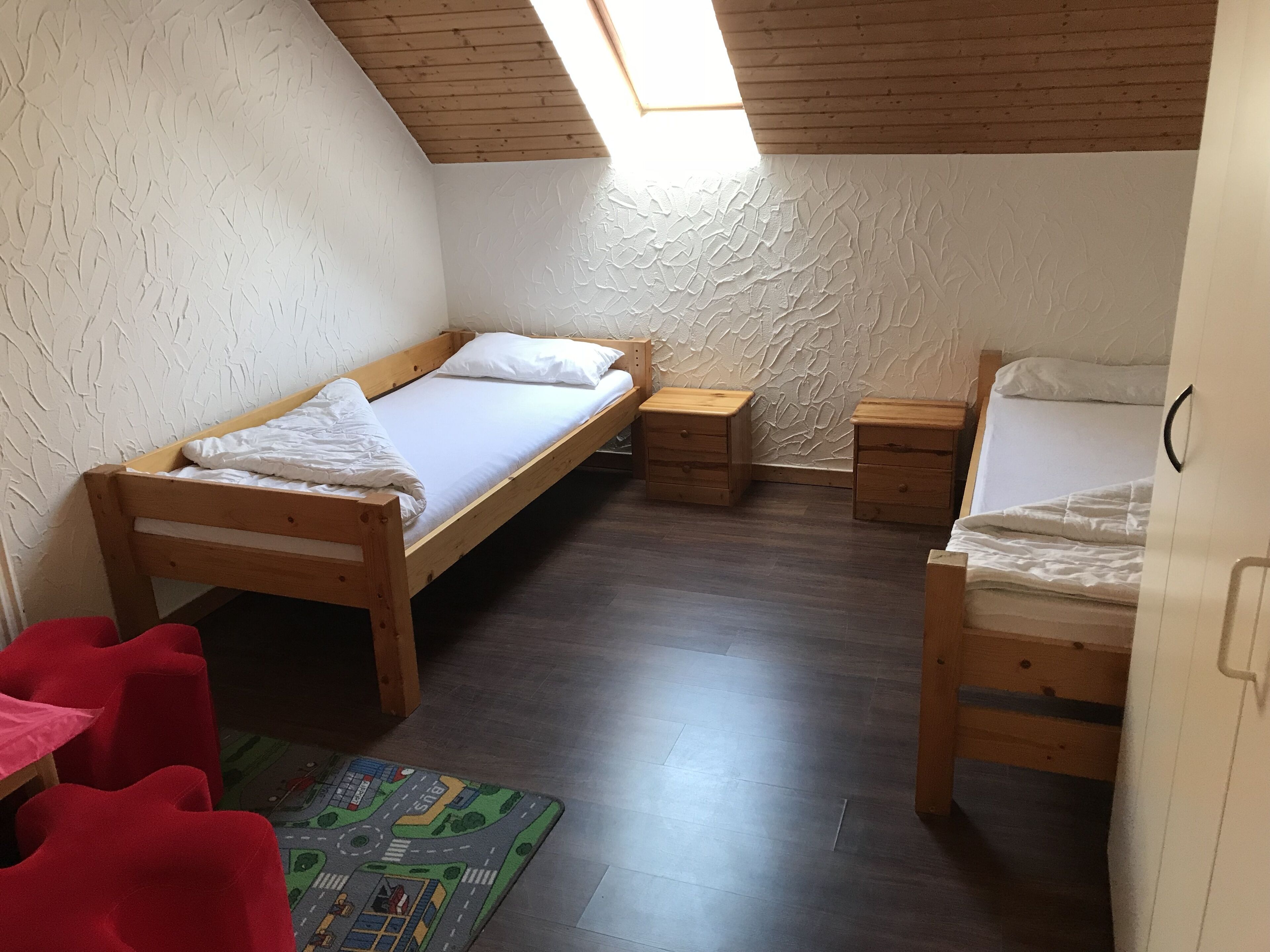 A small, cozy room with two wooden beds, a small table, and a red chair.
