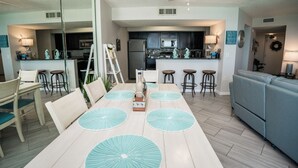 Une cuisine moderne avec des meubles de couleur sombre, un réfrigérateur et un four à micro-ondes. On y trouve un coin repas avec une table blanche, des sets de table bleus et des chaises. Un canapé bleu se trouve dans l’espace salon.
