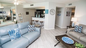 Un salon moderne avec un canapé bleu, une table basse en bois et un canapé gris. On aperçoit à l’arrière-plan un coin cuisine avec un réfrigérateur et une horloge au mur.