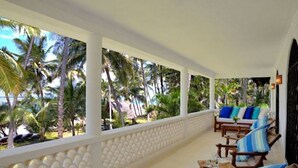 Een overdekt terras met een witte balustrade, houten meubels en uitzicht op palmbomen.