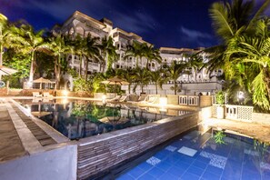 Vista nocturna de un hotel con piscina, palmeras y un edificio grande con balcones.