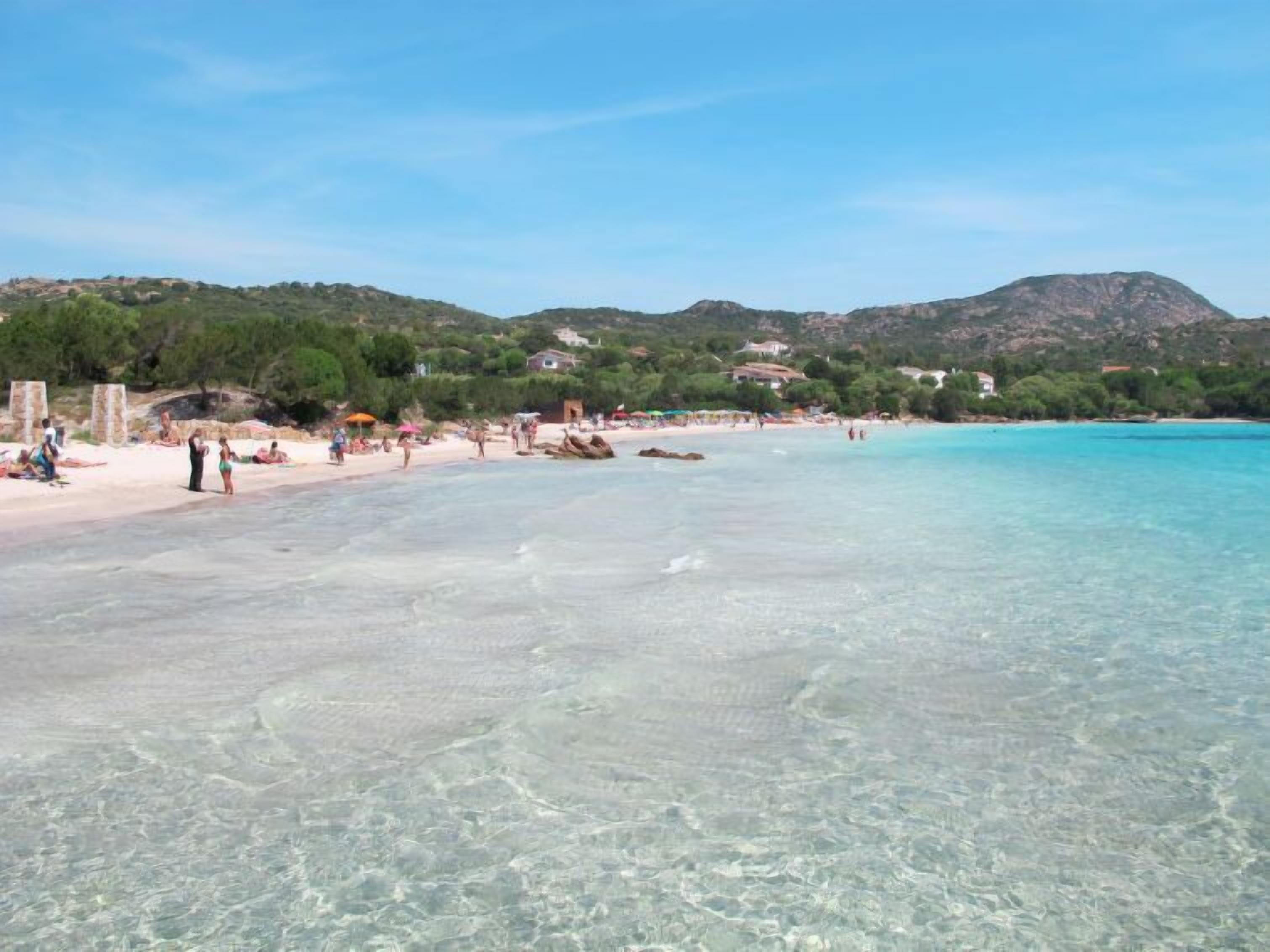 A beach with clear water, people, and buildings in the background.
