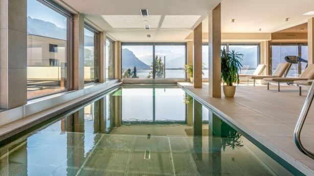 A modern indoor swimming pool with clear glass walls, a view of mountains, and lounge chairs.