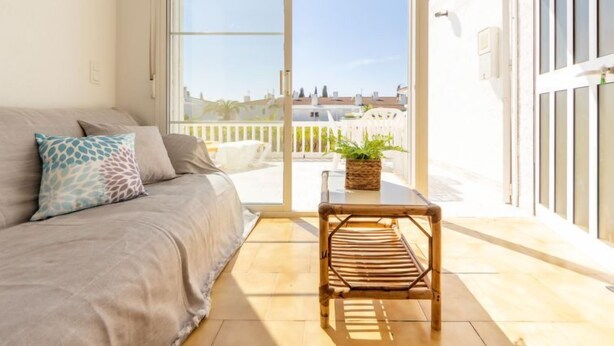 Un balcon avec un fauteuil en osier et un pot de fleurs, un canapé avec des coussins à motifs et une vue sur les immebles à travers de grandes portes vitrées.