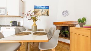 Une cuisine moderne avec des meubles blancs, une table à manger avec quatre chaises, un vase de fleurs, un tableau représentant une plage, une horloge et un banc en bois agrémenté d’une plante.