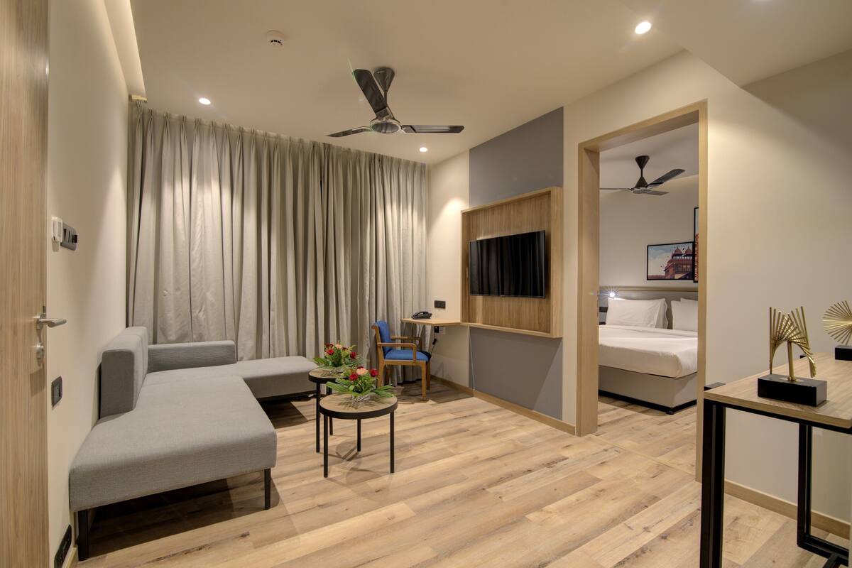 Ein modernes Hotelzimmer mit einer Couch, einem kleinen Tisch mit Blumen, einem an der Wand montierten Fernseher und einem Bett mit Kopfteil.