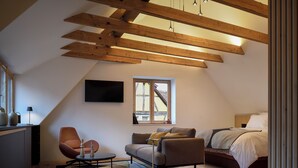 Een moderne loft slaapkamer met zichtbare houten balken, een groot raam, een bed, een bank en een bureau met stoel.